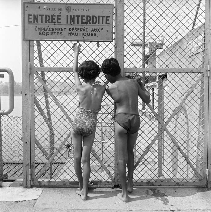 Deux enfants en maillot de bain observent le lac Léman à travers une grille